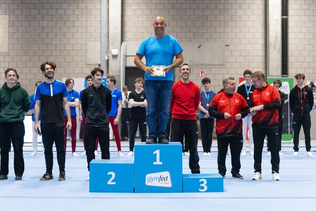 Male gymnast in blue shirt stands atop first-place podium holding trophy, surrounded by competitors in gymnastics hall