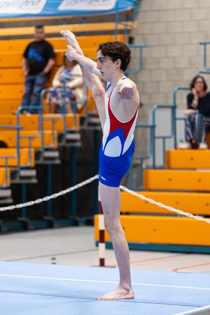 Young gymnast in red and blue leotard stands poised on floor mat, arms raised overhead in starting position