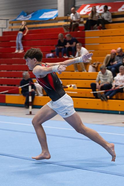 Male gymnast performs floor routine in lunge position with arms extended, competing barefoot on blue mat before spectators