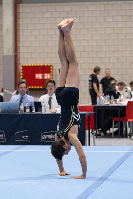 Male gymnast performs a handstand during floor routine as judges observe from nearby tables