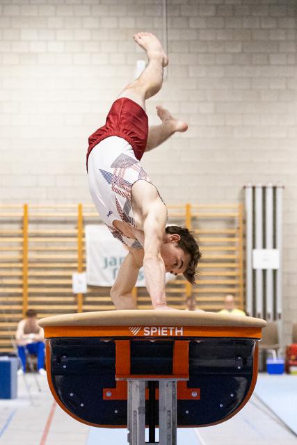 Male gymnast performs handstand on Spieth vault during training, legs pointed upward in gymnasium with wall bars visible