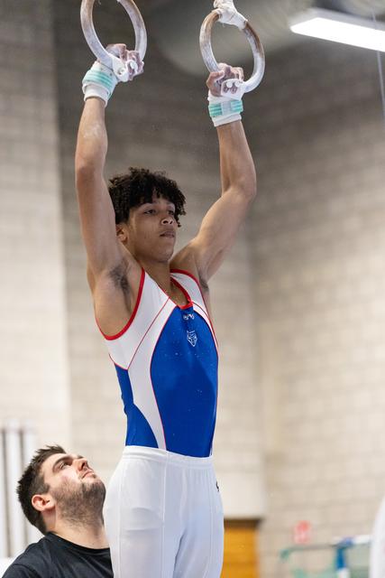 Young gymnast in blue and white leotard hangs from still rings while coach observes from below in indoor training facility