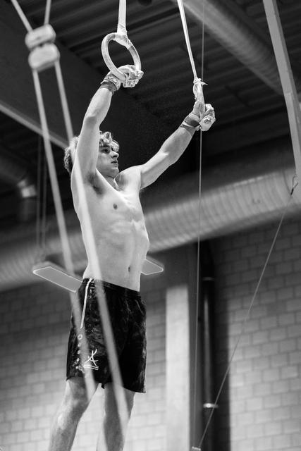 Shirtless male gymnast hanging from still rings in training facility, arms extended overhead, focused expression