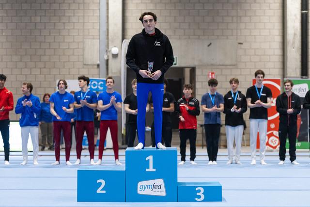 Male gymnast standing on first place podium holding trophy while competitors applaud in gymnasium background