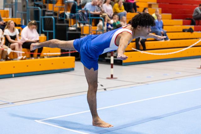 Male gymnast holds a horizontal arabesque position during floor routine, demonstrating strength and control in front of spectators