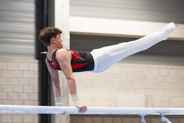 Male gymnast holds a horizontal position on parallel bars, demonstrating strength and control in white pants and red-black leotard