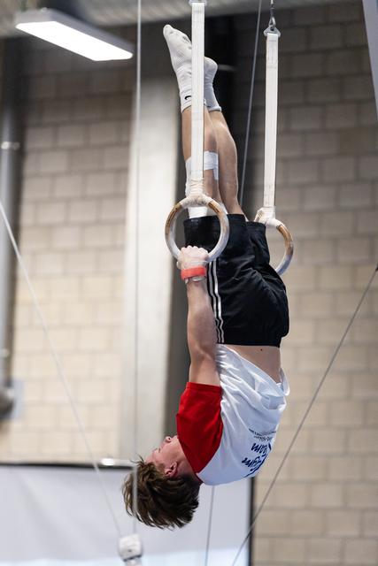 Male gymnast performing an inverted handstand on still rings, legs extended upward, in an indoor training facility