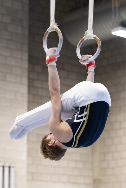 Male gymnast performs inverted tuck position on still rings with extended arms, showing strength and control indoors