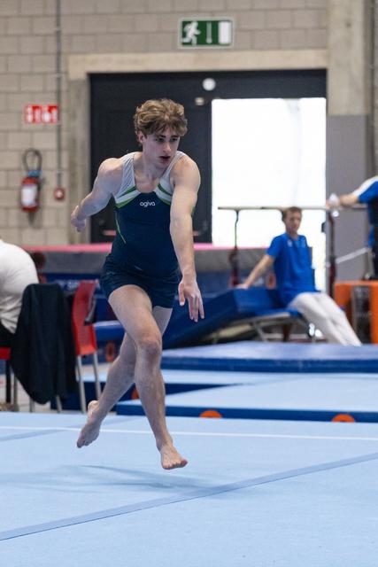 Young gymnast landing on floor exercise mat with intense concentration, wearing dark leotard in training facility