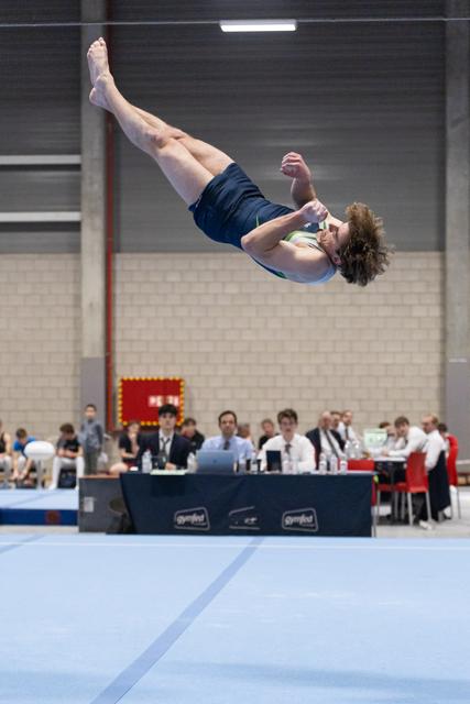 Gymnast performs a tucked backflip during floor routine as judges observe in background at indoor gymnastics event