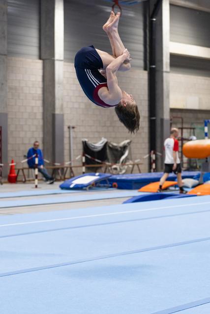 Gymnast performs a tight backflip mid-air above blue training mats in indoor gymnastics facility