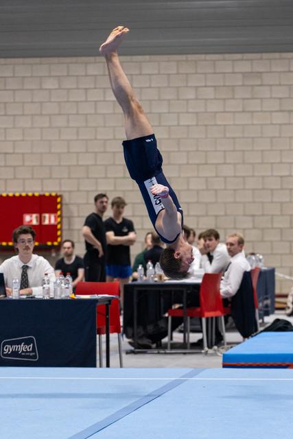 Male gymnast performs inverted backflip on floor exercise while judges observe from tables in indoor venue