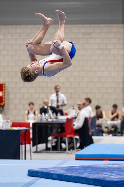 Male gymnast performs a tucked backflip mid-air on trampoline, captured in sharp detail against blurred judges' table