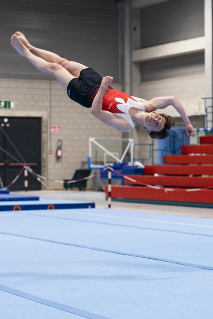 Gymnast executing a backflip during floor exercise in training facility, body fully inverted mid-air above blue mat