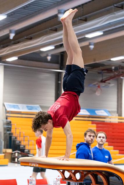Male gymnast executing a handstand on vault apparatus while two coaches in blue jackets observe from behind