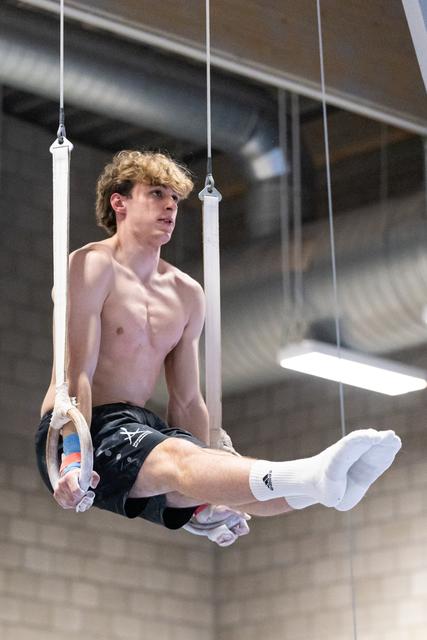Shirtless male gymnast holds an L-sit position on still rings, displaying strength and control in an indoor training facility