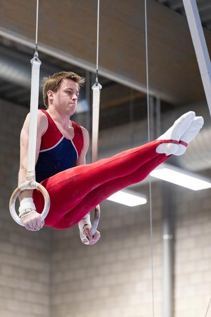 Male gymnast performing a horizontal hold on rings, legs extended in red pants, displaying strength and concentration