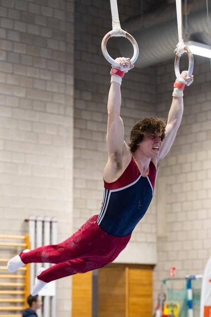 Young male gymnast performing on rings with extended body position, wearing red and blue leotard in indoor training facility