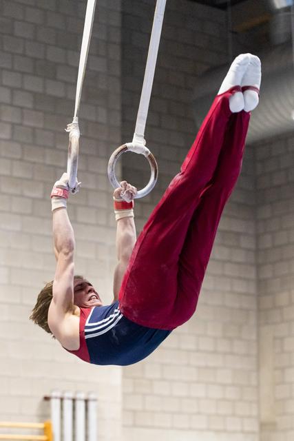 Male gymnast performs an L-sit position on rings, suspended in air with intense focus in an indoor training facility