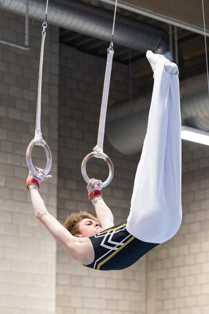 Gymnast performing on rings in navy leotard and white pants, arms extended upward in an indoor training facility