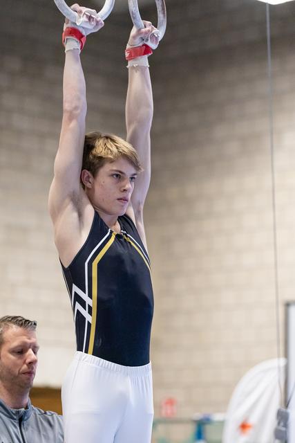 Young male gymnast performing a hang position on still rings while coach observes intently during training session