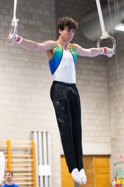 Young male gymnast holds iron cross position on rings, wearing colorful leotard with white pants and grips in gymnasium
