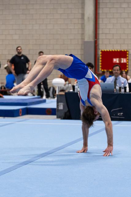 Gymnast executing a dramatic backbend during floor routine, arching backward with hands planted on blue mat