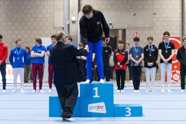 Athlete in blue pants stands on first place podium receiving medal from official as competitors watch in background