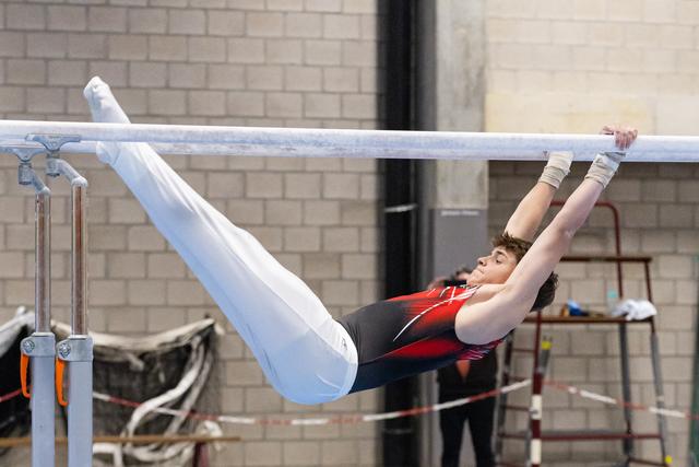 Gymnast in red and black leotard performs horizontal bar routine with extended body position and white pants on legs