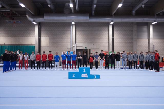Teams lined up on podium positions at gymnastics venue, athletes in matching team uniforms standing together after awards ceremony