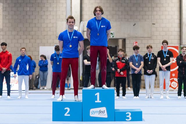 Two gymnasts stand on a medal podium wearing blue shirts and burgundy pants, with other athletes watching in the background