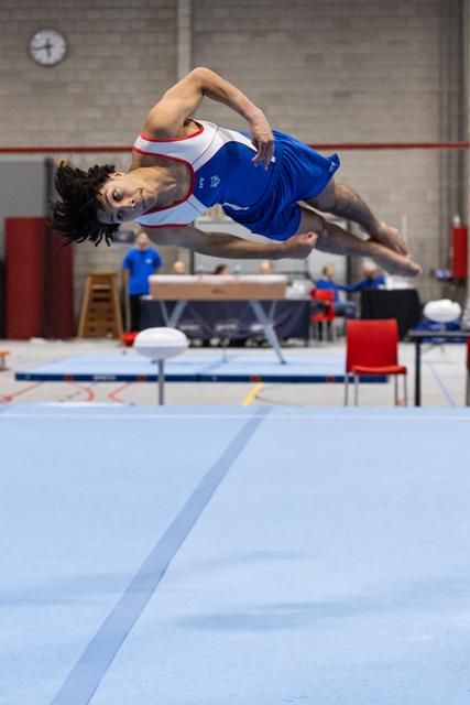 Male gymnast performs a dynamic twisting jump during floor routine, body extended mid-air in training facility