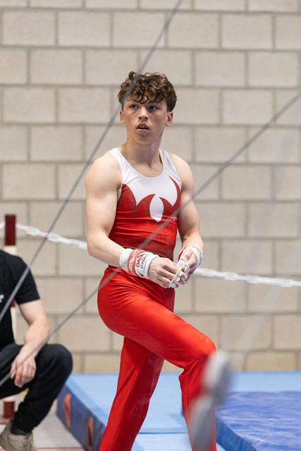 Male gymnast in red and white uniform chalking hands before high bar routine, intense focus visible on face