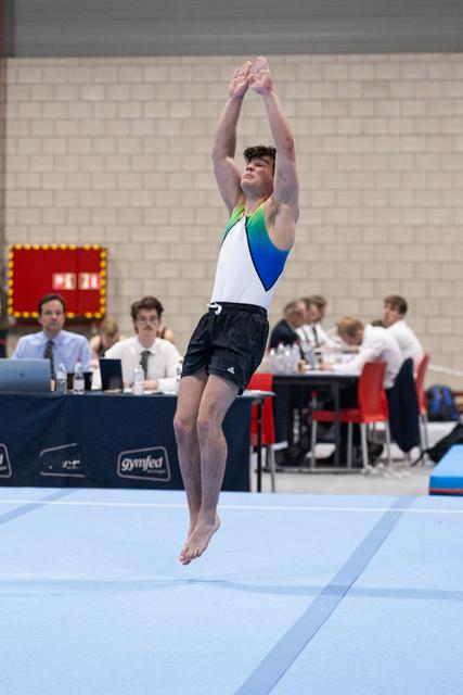 Male gymnast performing a vertical jump with arms raised overhead during floor exercise routine at indoor competition