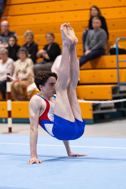 Male gymnast performs an L-support position during floor exercise routine, demonstrating strength and control before spectators
