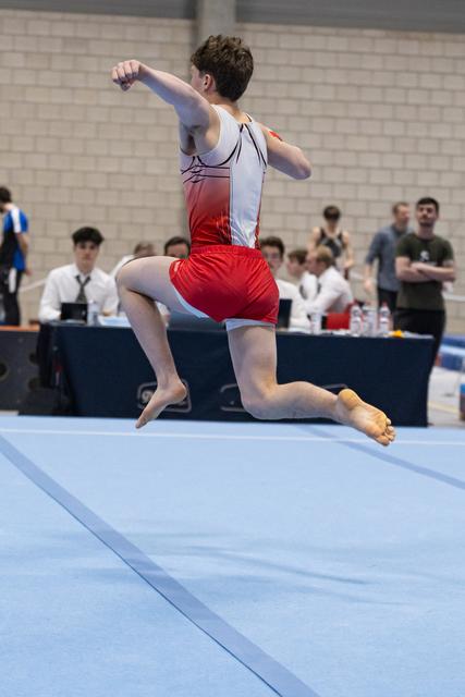 Male gymnast mid-leap during floor routine, arms positioned dynamically, performing before judges and spectators