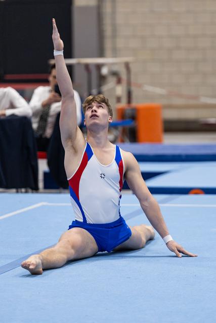 Male gymnast in white and blue leotard performs an elegant floor routine finishing pose, arm extended upward, looking skyward