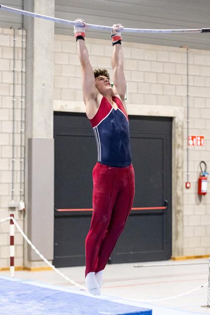 Male gymnast performing a high bar routine with intense concentration, wearing red and blue competitive attire in an indoor gym.