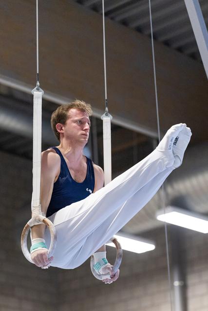 Male gymnast performs on rings with extended body position, wearing navy leotard and white pants in indoor facility