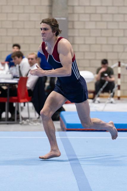 Male gymnast in navy and maroon leotard running barefoot across blue floor mat during routine, focused expression