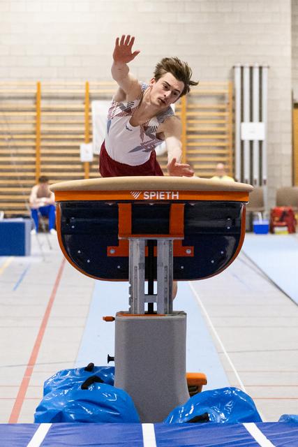 Male gymnast mid-flight over Spieth vault, arms extended upward, hair airborne, in indoor training facility