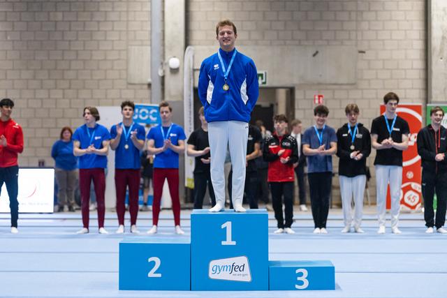Winner in blue tracksuit stands atop first-place podium with medal, beaming with pride as fellow competitors stand on lower positions