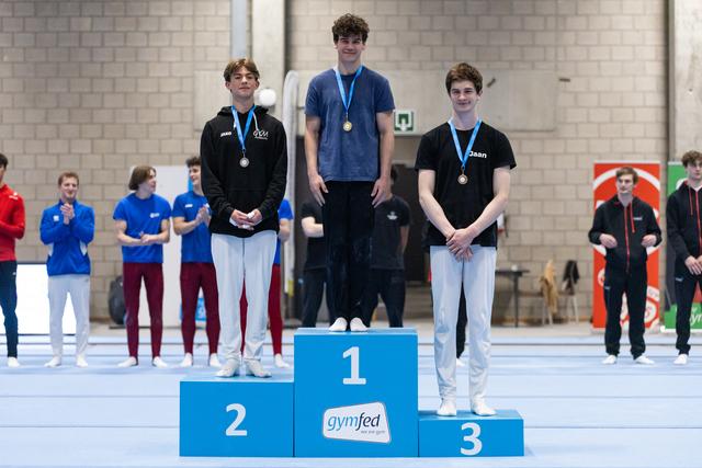 Three young male gymnasts stand on award podium wearing medals, with first place holding gold and teammates in silver and bronze positions