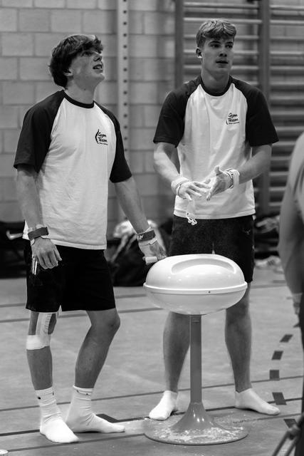 Two young gymnasts in matching raglan shirts share a laugh while standing beside a mushroom training apparatus in the gym