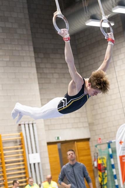 Young gymnast performing an iron cross position on rings while coach watches from below in indoor training facility