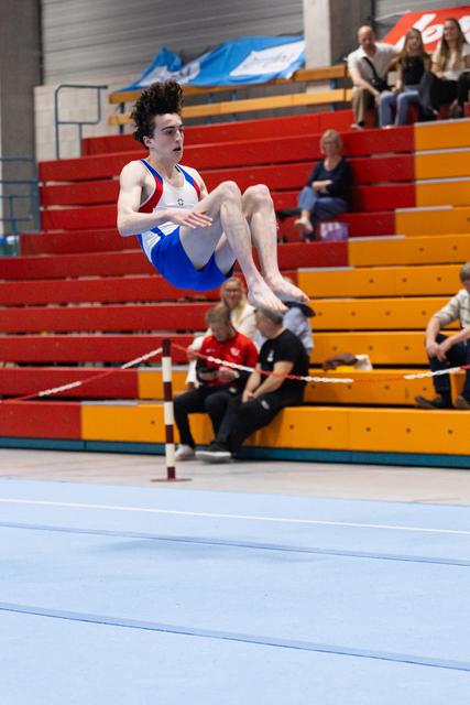 Young male gymnast performs a tucked flip mid-air during floor exercise routine with spectators in bleachers behind