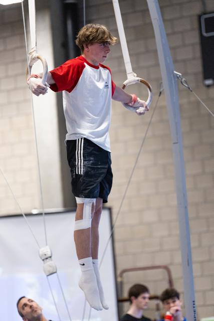 Young gymnast in red and white shirt performing on rings, holding position with focused concentration in training facility