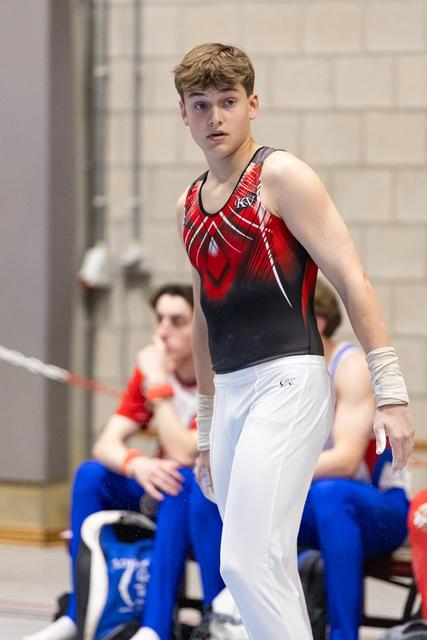 Young gymnast in red and navy leotard stands alert, watching intently while teammates sit in background