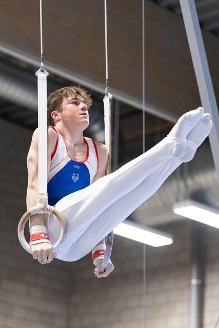 Male gymnast performing rings routine with extended legs, showing intense focus and strength in white pants and red-blue leotard