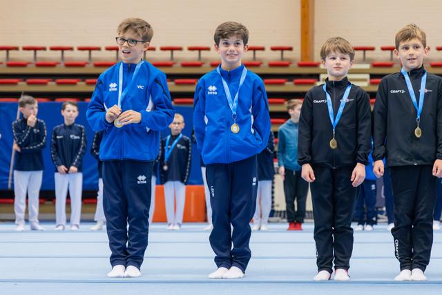 Four young gymnasts stand proudly on the podium, wearing gold and silver medals, smiling in blue and black tracksuits.